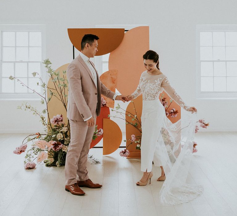 Bride wears white trousers and long sheered detailed top as she smiles and dances with groom in linen light suit at their asian wedding 