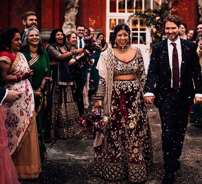 Guests cheer & throw confetti as Bride in burgundy wedding dress and Groom matches in burgundy tie 