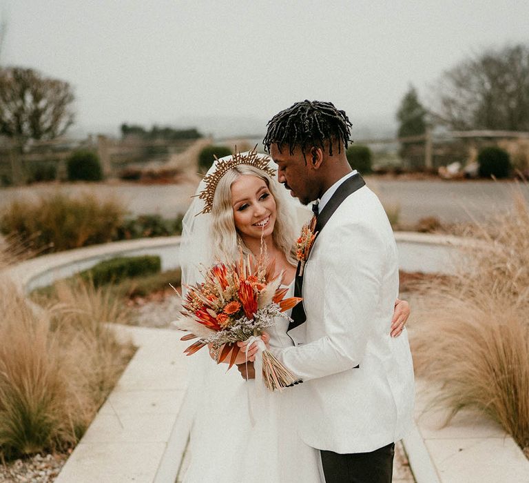 Groom in white tuxedo embraces his bride holding orange bouquet