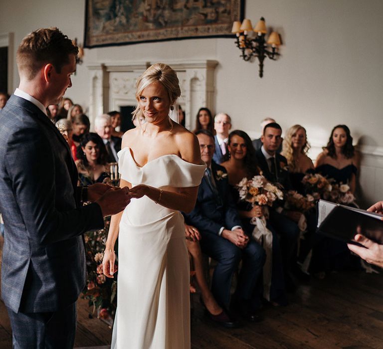 Bride in sweetheart off the shoulder wedding dress and blonde updo smiles at groom who places ring on her finger