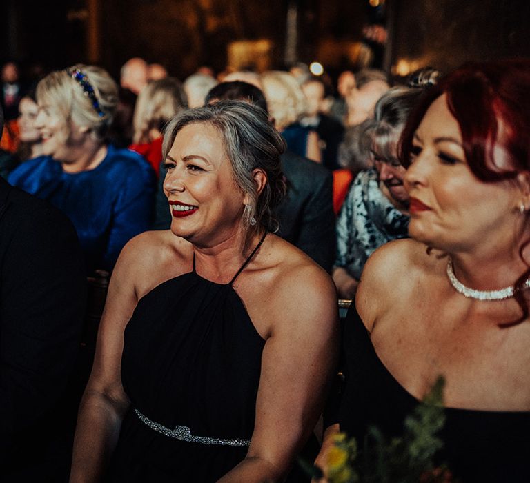 Wedding guests in black dresses and bold red lips smile as the grooms get married 