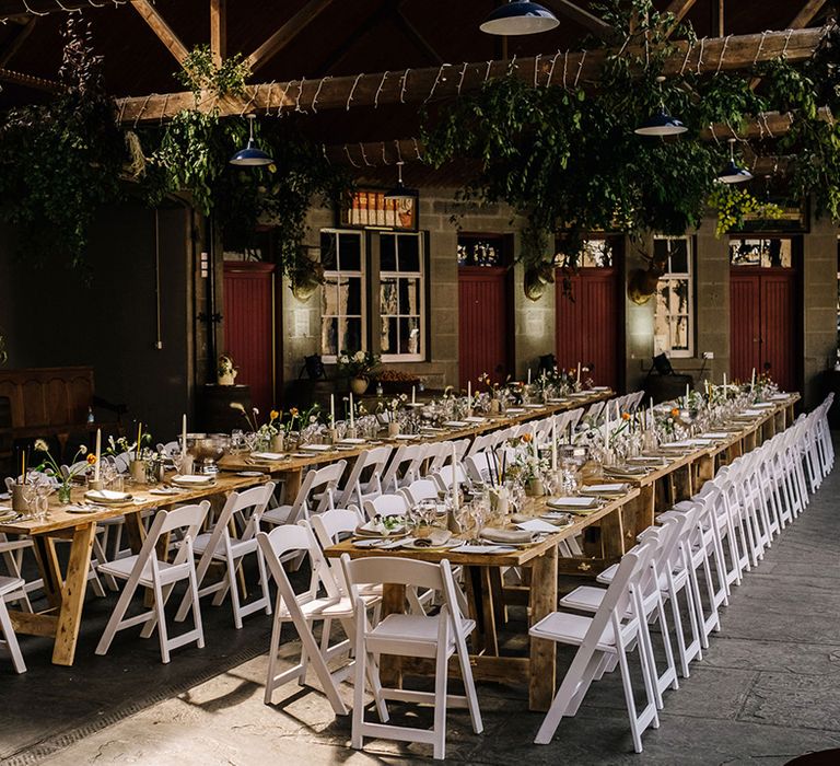 Wedding reception layout with long wooden banquet tables and white wooden chairs with colourful flowers and foliage above 