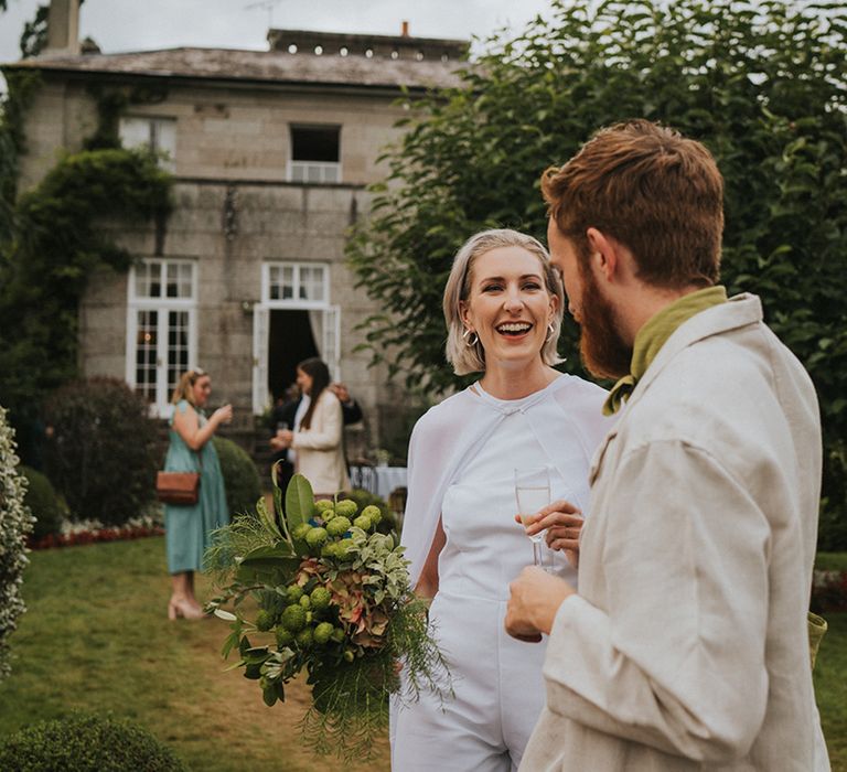 Bride in jumpsuit with cape holding leaf wedding bouquet speaks to wedding guests 