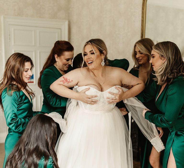 Bridesmaids in emerald green v-neck long sleeve dresses with a front slit help the bride into her custom wedding dress