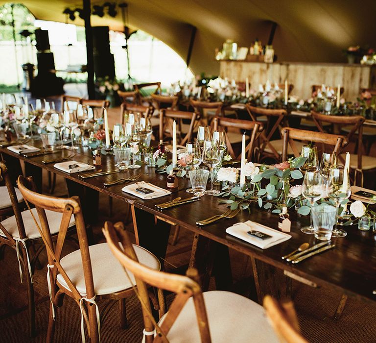 Dark wooden banquet tables and wooden chairs with light pink and green foliage runner with white candles 