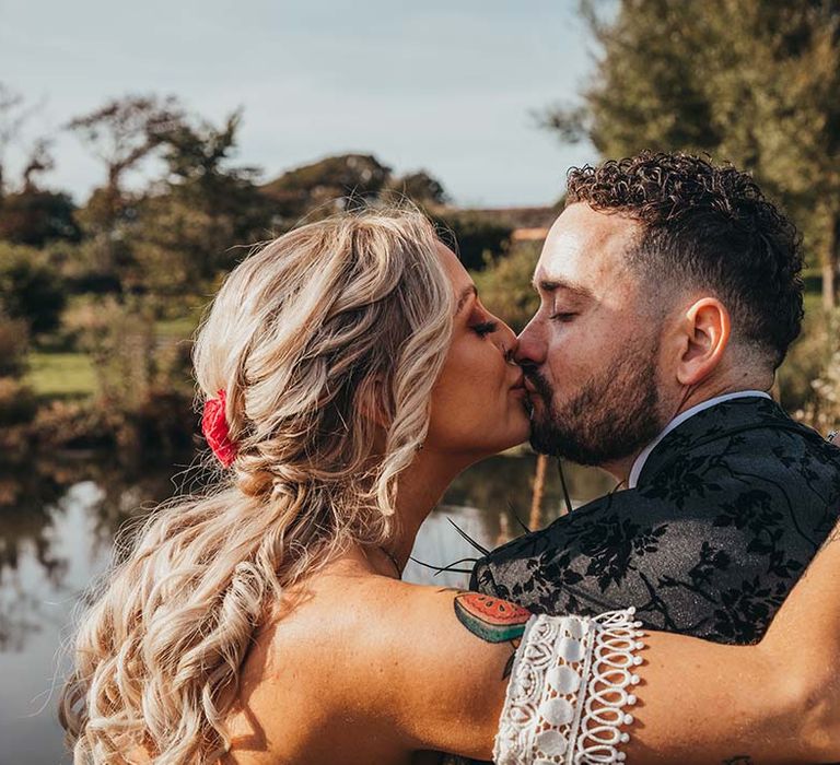 Bride with braided curly blonde hair with a red flower accessory kisses groom in black patterned suit 