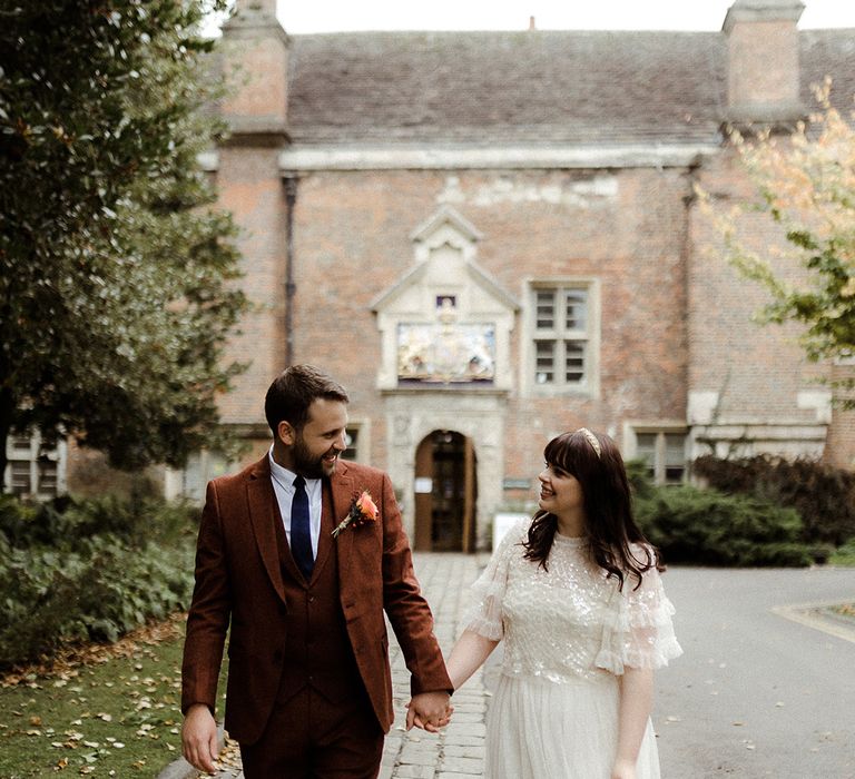 Groom in brown three piece suit and blue tie holds hands with bride in Needle and Thread bridal separates and gold headband and shoes