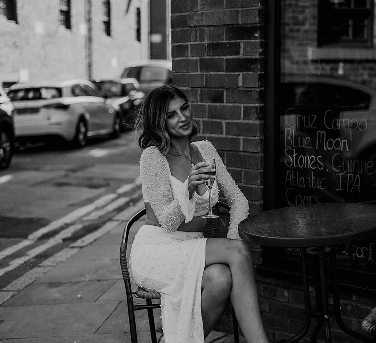 Bride in pearl embellished two piece with white feather wedding shoes holding champagne flute outside venue 