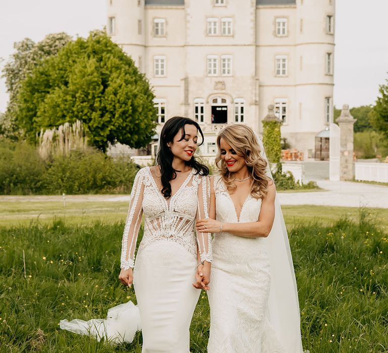 Bride in long sleeve illusion lace wedding dress and bride in v-neck lace wedding dress walk holding hands together 