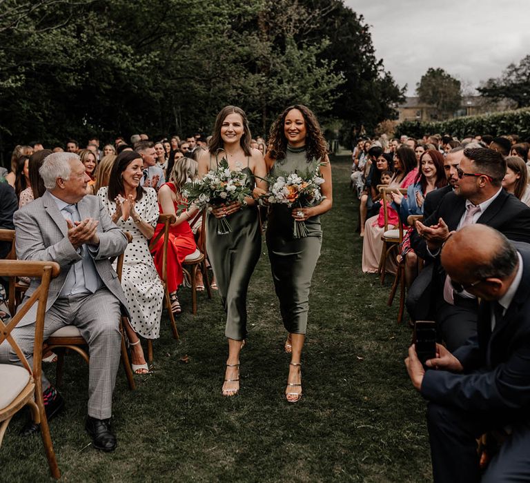 Bridesmaids in satin sage green bridesmaid dresses walking down the aisle at an outdoor back garden wedding 