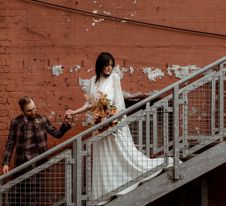 Groom leads bride down industrial staircase on their wedding day for couples portraits 