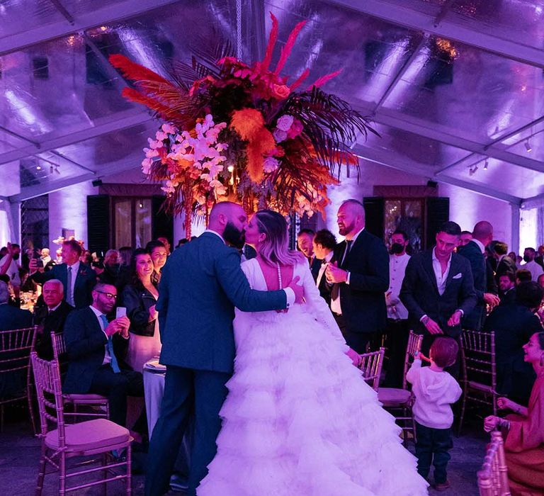 Bride & groom kiss in front wedding guests as brightly coloured floral installation hangs above them 