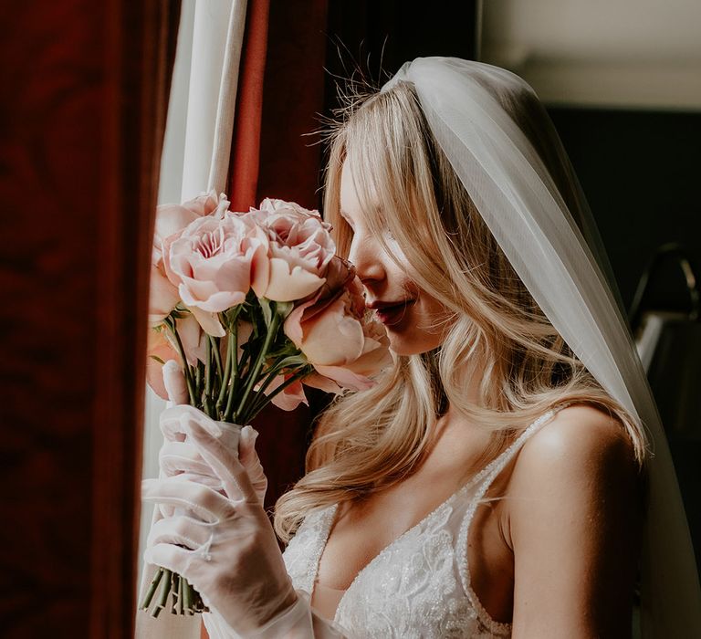 Bride in sparkly wedding dress by Made With Love with tulle bridal gloves smelling her pink flower round bouquet