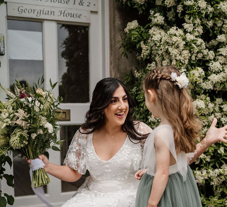 Bride kneels in boho wedding dress whilst speaking to flower girl who wears white and sage green dress with her brown hair in a plait tied with white ribbon