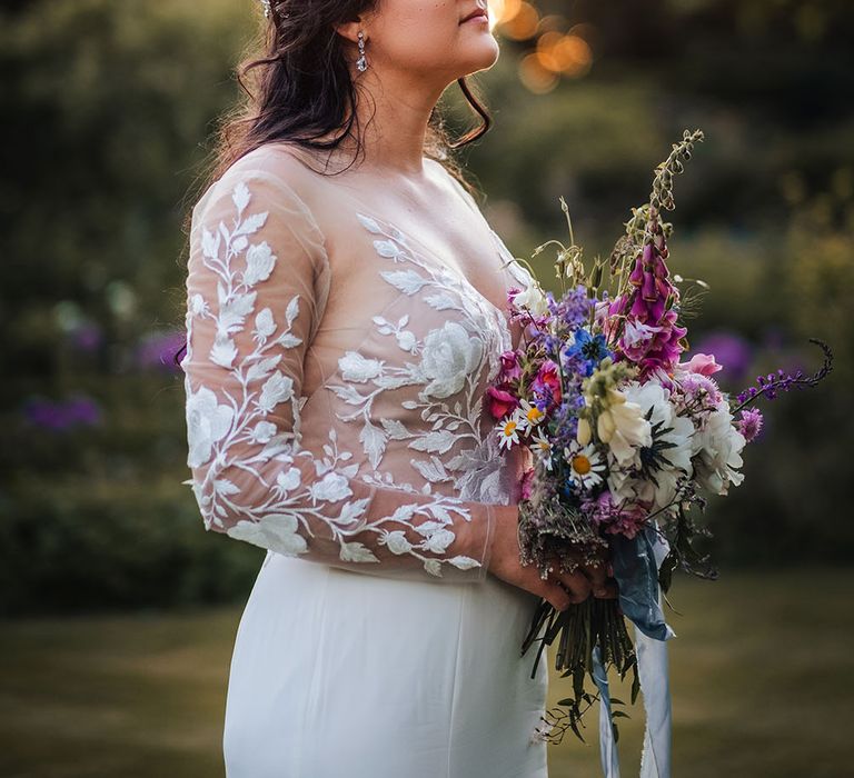 Bride wears her dark hair pulled back into loose curls complete with hair accessory and holds tropical bouquet whilst wearing sheer lace embellished wedding dress