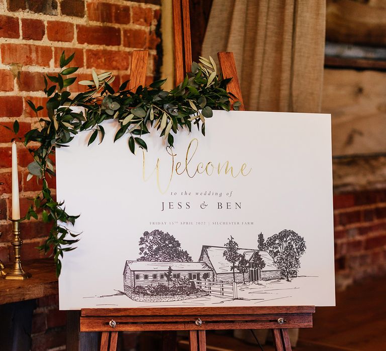 Black, white and gold welcome wedding sign with a sketch of the wedding venue and decorated with green foliage 