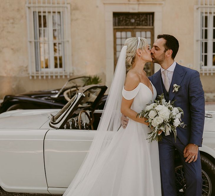 Bride & groom kiss in front of vintage car outside the Chateau de Robernier after wedding ceremony in the South of France