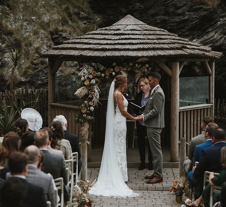 Bride & groom stand in front of beach styled hut surrounded by champagne and rust coloured florals for outdoor ceremony