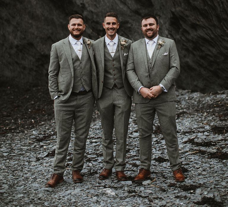 Groom stands with his groomsmen who all wear three piece suits with floral buttonholes and neutral ties 