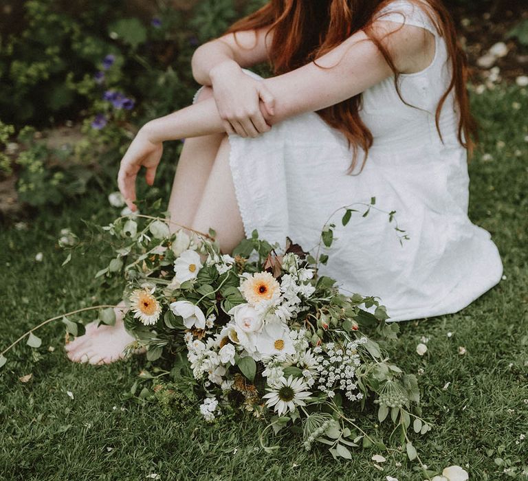 Beautiful bride with long hair and natural makeup sitting on the grass with her green, white and yellow flower and foliage wedding bouquet 