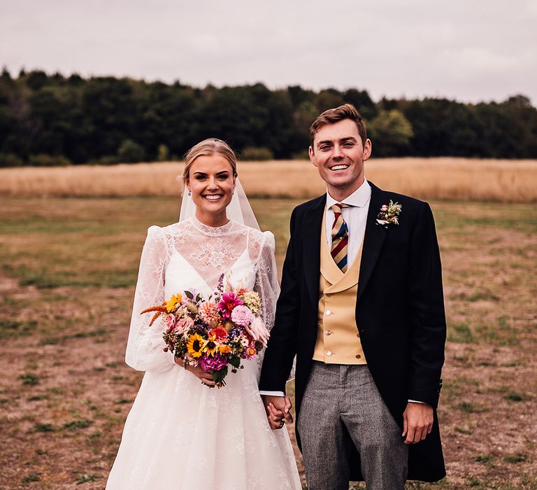 Groom in grey trousers, yellow waistcoat, striped tie and black morning suit stands holding hands with the bride in a long sleeve delicate lace wedding dress