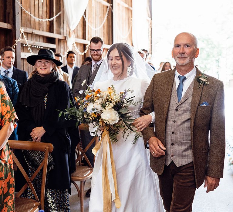 Father of the bride in a brown suit with a dark blue tie walks the bride down the aisle in a vintage lace wedding dress with a guest getting emotional 