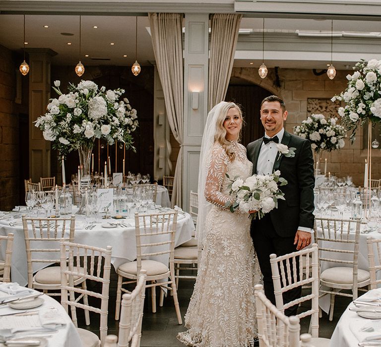 Bride wearing lace wedding dress with high neck and long sleeves stands with her groom in black tie