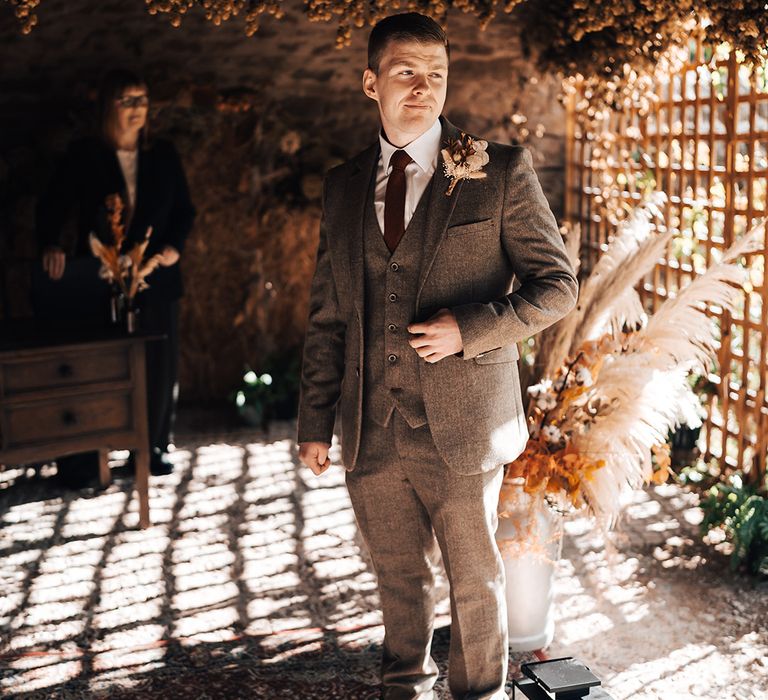 Groom wears brown three piece suit with burnt orange tie and dried floral buttonhole