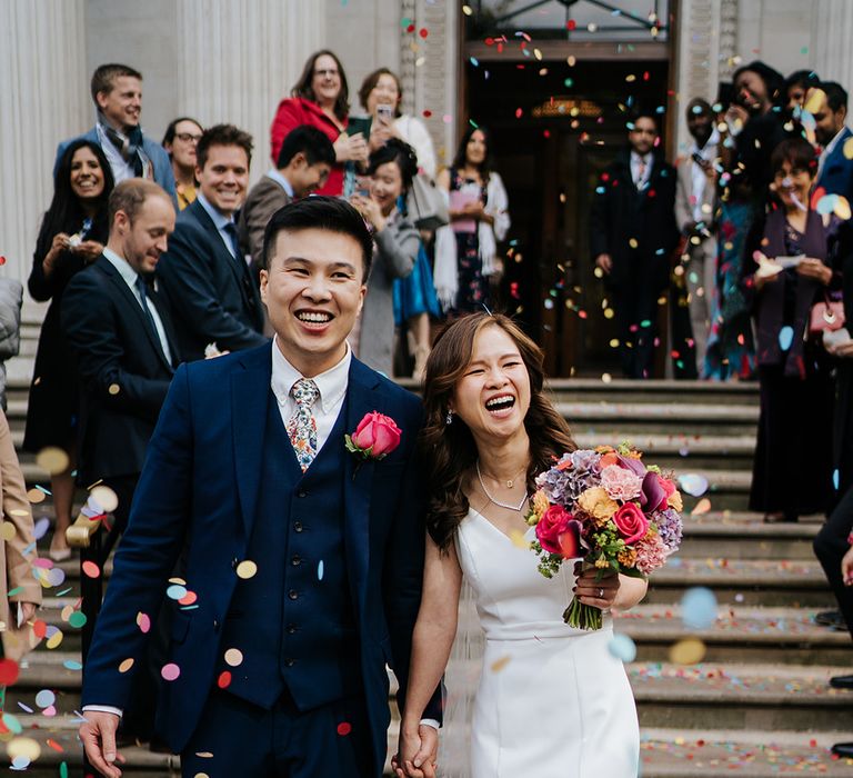 Bride holds bright floral bouquet and walks with her groom through colourful confetti exit at the Old Marylebone Town Hall