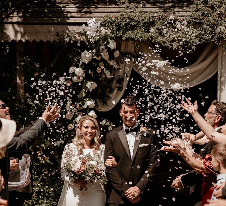 Bride in a long sleeve fitted wedding dress walking with the groom in black tie have their confetti exit moment 