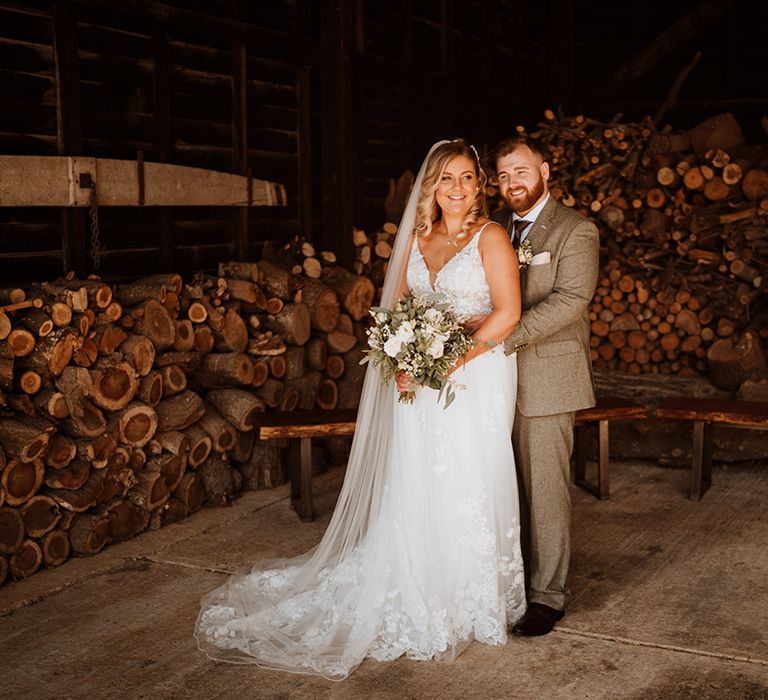 The groom in a grey tweed suit embraces the bride in a lace wedding dress from behind for their rustic wedding 