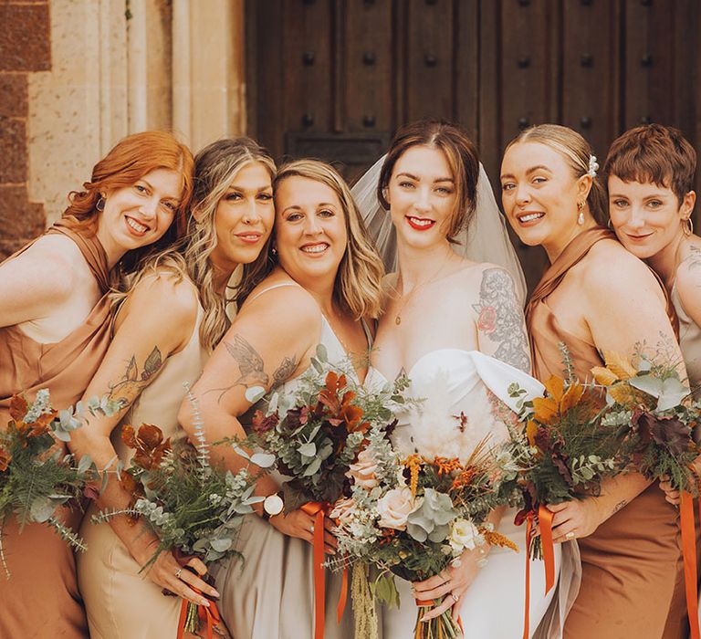 Bride with tattoos stands with her bridesmaids in Autumnal bridesmaid dresses and bouquets tied with orange silk ribbon 