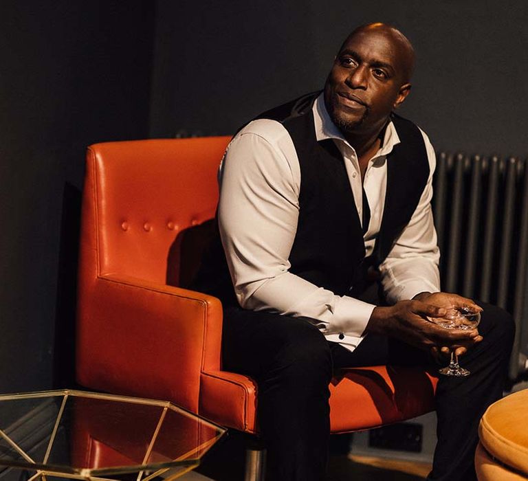 Groom in black waistcoat and suit trousers with slightly opened white shirt and glass of Champagne sitting on red leather chair 