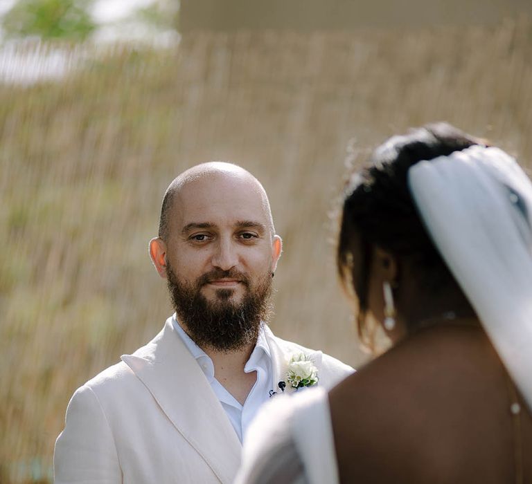 Groom in cream suit smiling at the Bride