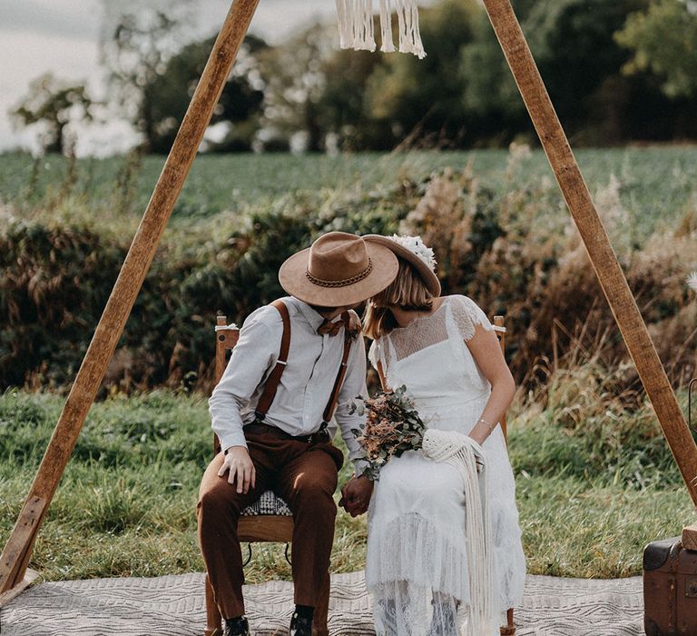 Wooden arch decor with handmade macrame decor with the bride and groom seated on wooden chairs wearing stylish hats