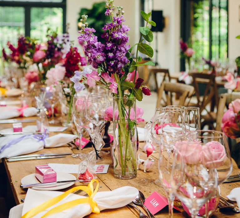 Neutral wedding tablescape with pops of pink, purple, yellow and fuchsia - glass bottles with large purple floral arrangements, white napkins with yellow and lilac ribbon, and fuchsia place names