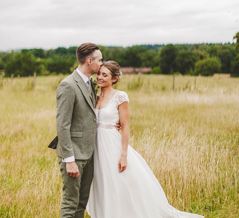 Groom in grey suit kisses boho Bride in a field during outdoor wedding photography