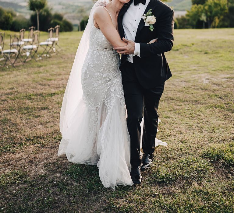 Bride in plunge lace wedding dress and trumpet styled ruffled skirt hugs her groom in black-tie 