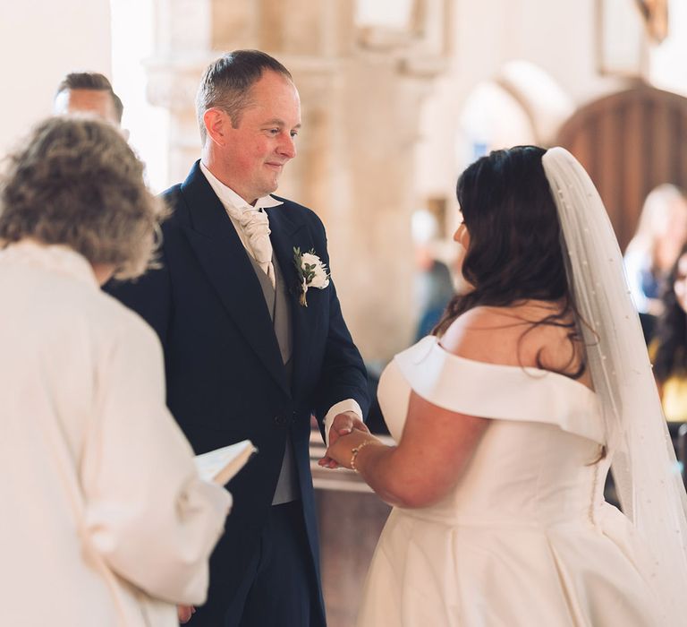 Groom in traditional morning suit in a navy shade with a white flower buttonhole holding hands with the bride in an off the shoulder wedding dress for their church ceremony 