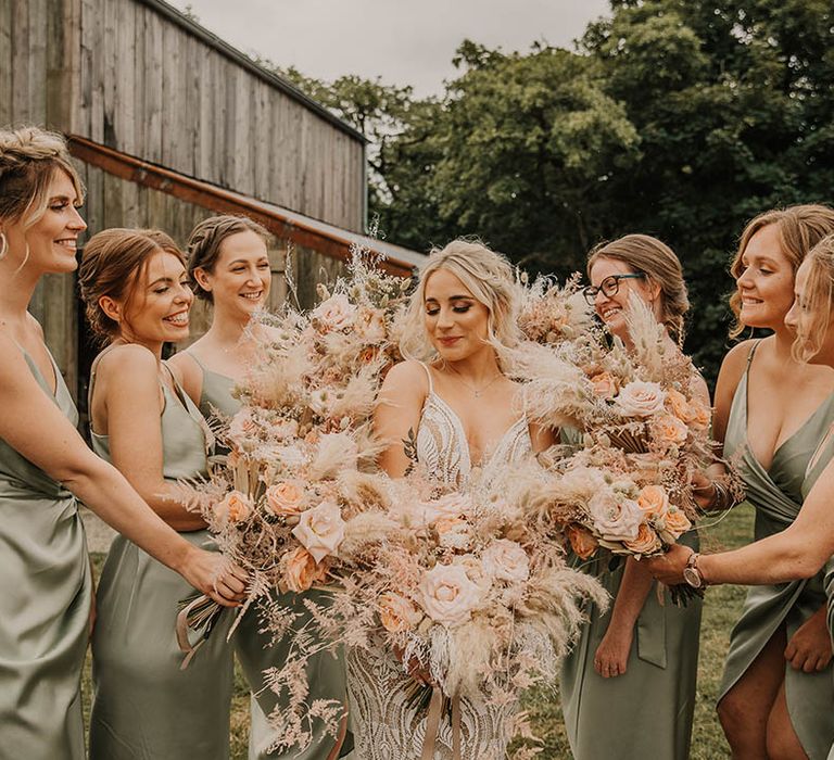 The bridal party in sage green satin bridesmaid dresses hold their bouquets around the bride in a fitted lace gown 