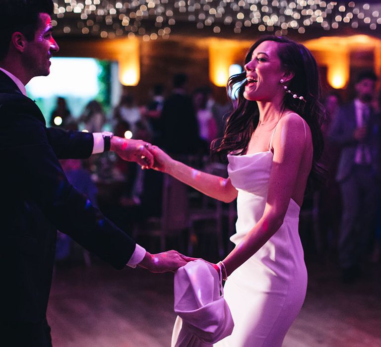 Groom in dark navy suit sharing the first dance with the bride in a satin gown as pink lights shine on them 