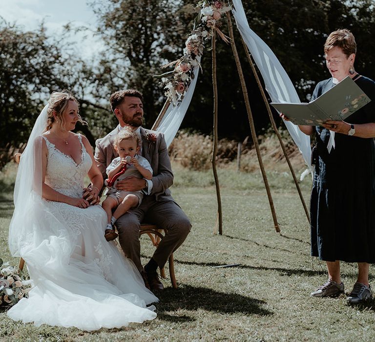 Bride & groom sit with their baby during outdoor wedding ceremony at The Apple Orchard