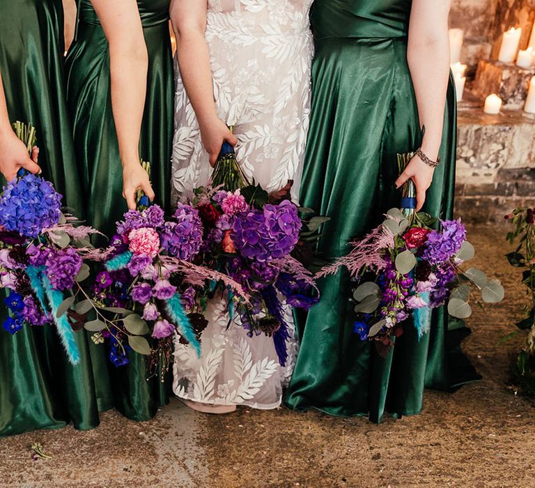 Bridesmaids in forest green satin bridesmaid dresses and bride in 3d appliqué flowing sheer bridal gown holding bright purple bridal bouquets with purple hydrangeas, pink garden roses, purple orchids, lilac carnations, dried flowers and peacock blue feathers 