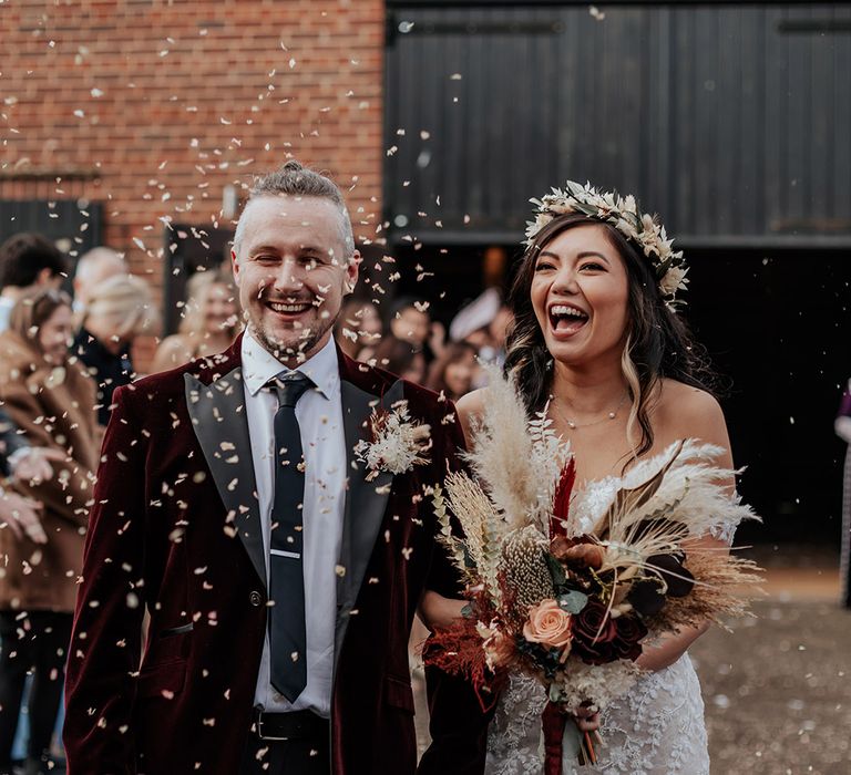 Groom in velvet suit jacket laughing as he walks along with the bride in an off the shoulder lace wedding dress with boho flower crown having confetti exit 