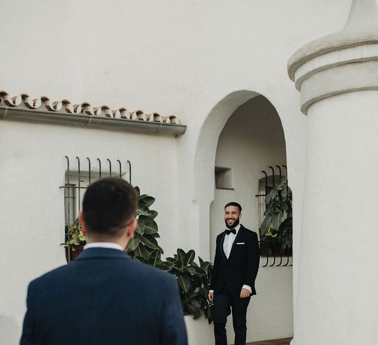 Groom in a navy suit waiting for his groom in a black tuxedo at their first look