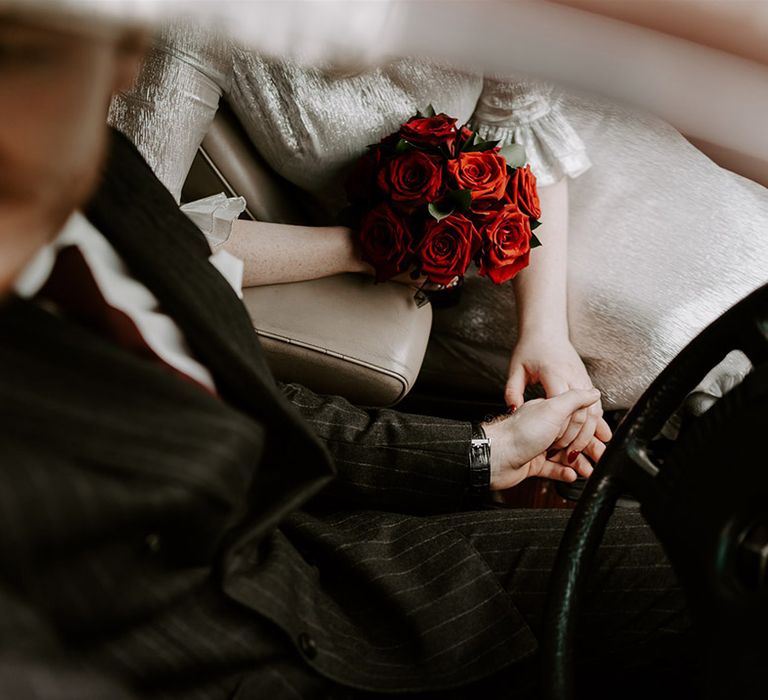 The groom in a pinstriped wedding suit driving the wedding car holding hands with the bride in a metallic wedding dress holding the red rose bouquet 