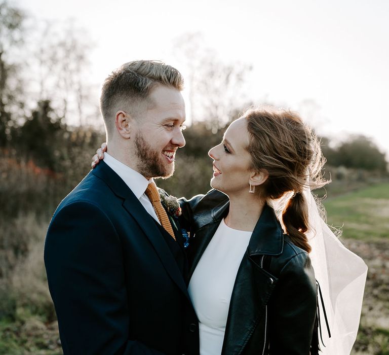 Bride in tassel leather jacket with wedding veil embracing the groom in a navy suit with a yellow tie 