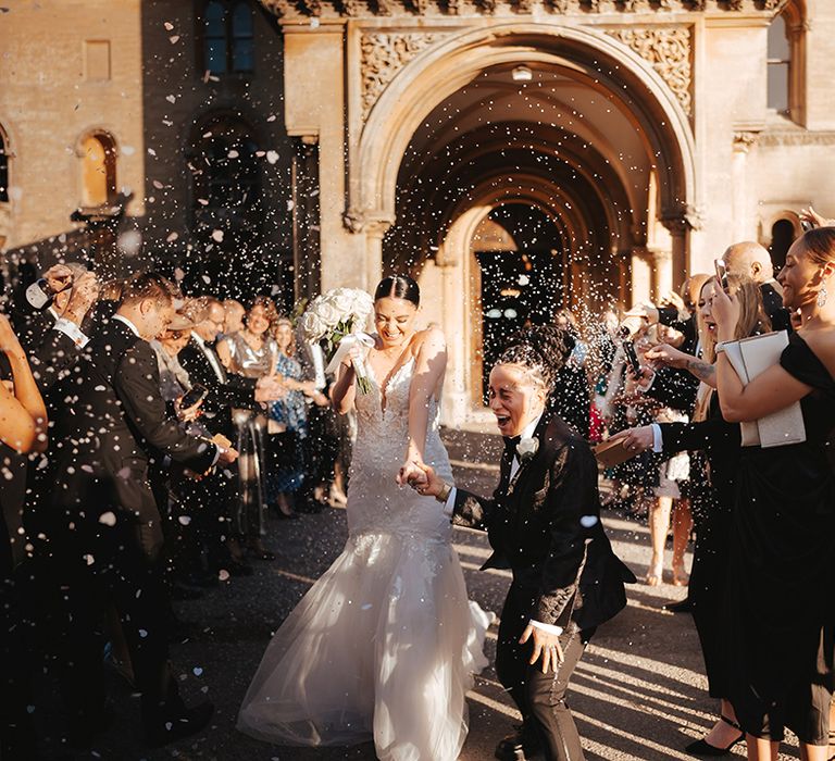 Bride in lace sleeveless v neck Enzoani wedding dress with tulle finish and church length veil holding hands with Bride in black snakeskin print tuxedo with black bowtie and white rose boutonniere holding white garden rose bridal bouquet doing white petal confetti exit at wedding