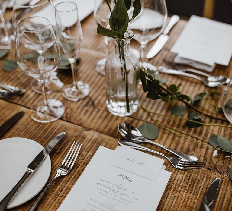 Black and white wedding menu on wooden table with white plates, silver cutlery and bud vases with pink roses 