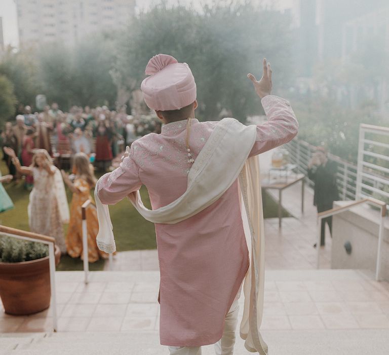 Groom in light pink sherwani, light pink shoes and light pink turban and cream trousers doing traditional Indian dance at wedding